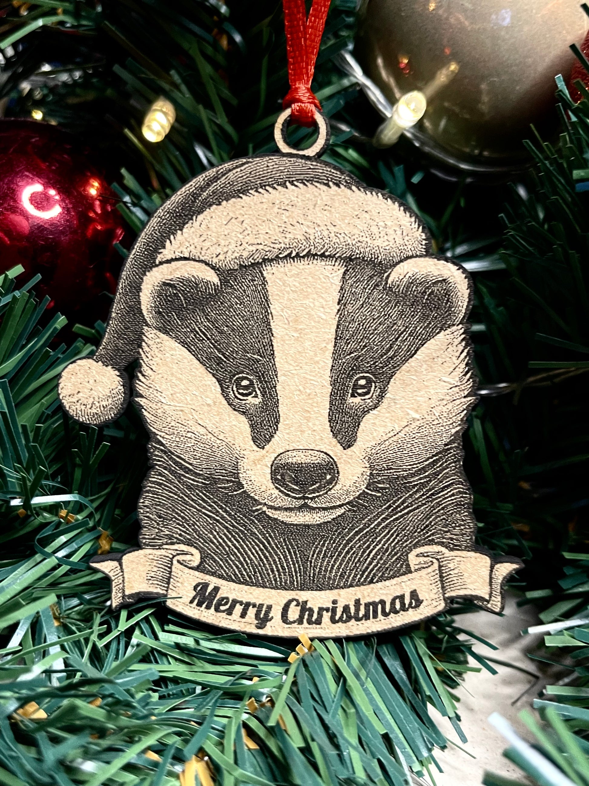A wooden badger tree ornament with a Santa hat and a banner that reads 'Merry Christmas', hanging among Christmas tree branches.